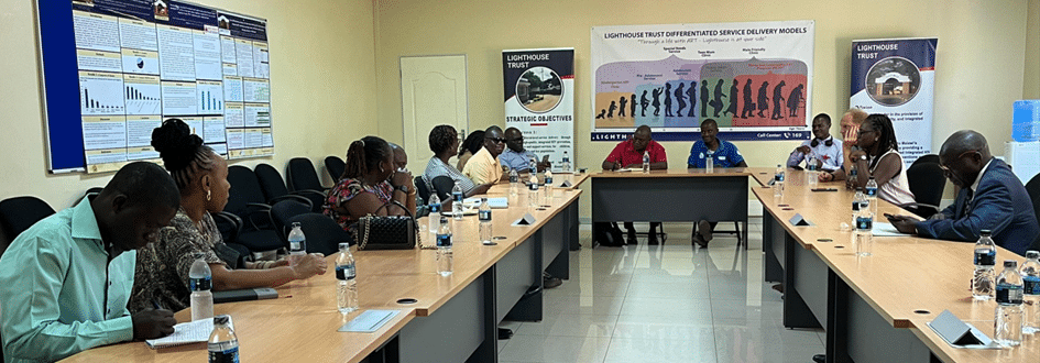 A group of people seated around a conference table in a room with posters on the walls. The posters display charts and logos. Two banners stand at the front of the room next to a projector screen. Bottles of water are on the table.