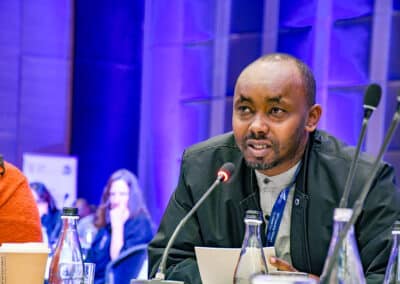 A man speaks into a microphone at a conference table, with bottles and papers in front of him. The background is blue-lit, and other attendees are visible, seated and listening.