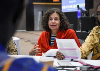 A woman with curly hair, wearing a red shawl and striped shirt, speaks while holding papers at a table with others during a meeting or workshop. A coffee cup and documents are on the table. A screen is visible in the background.