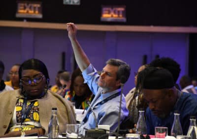 A man raises his hand during a conference, surrounded by other attendees who are seated and focused. Bottles and microphones are on the table, and "EXIT" signs are visible in the background.