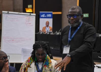 A man wearing glasses and a lanyard speaks while standing beside a seated woman at a conference. Behind them is a flip chart with handwritten notes and a conference banner featuring a smiling person.