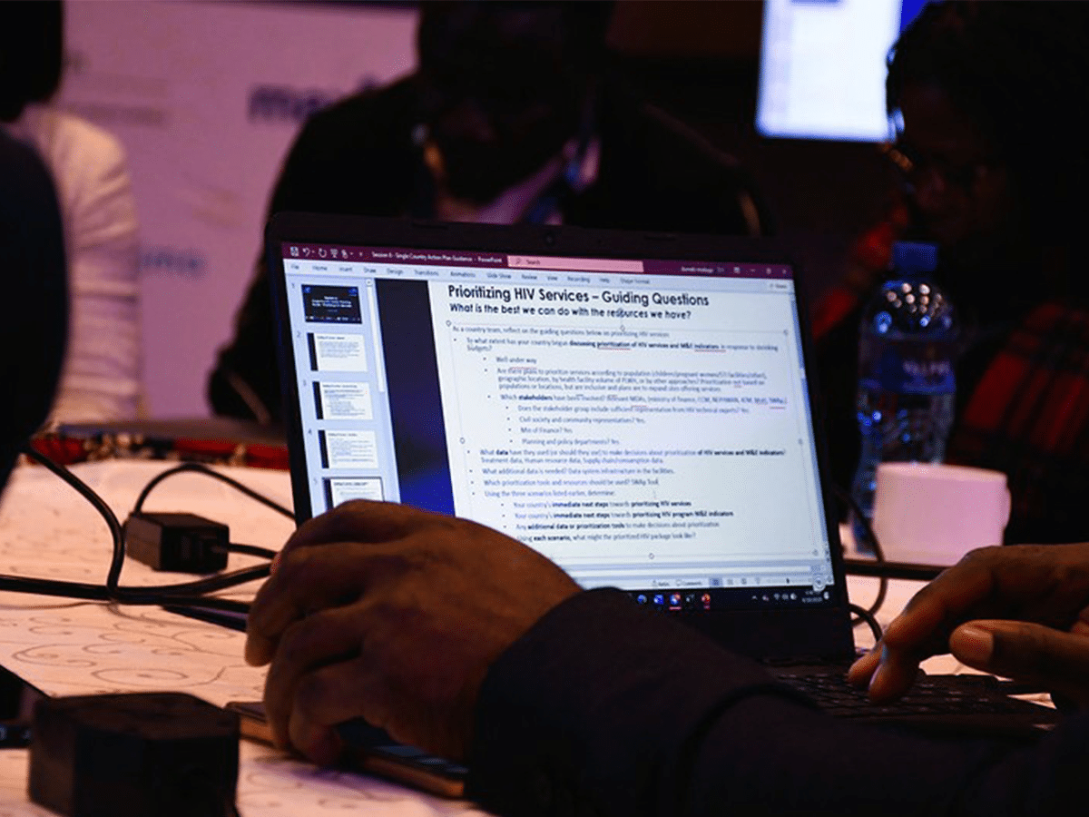 cquin pivot cover A person uses a laptop displaying a presentation slide titled "Prioritizing HIV Services – Guiding Questions" at a table with other people, papers, and a water bottle visible in the background.