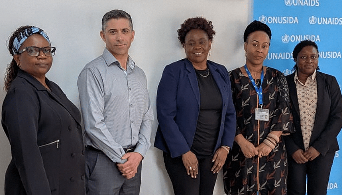 Five people stand together indoors, posing for a group photo in front of a blue banner with the UNAIDS logo. Four women and one man are dressed in professional attire and smiling at the camera.
