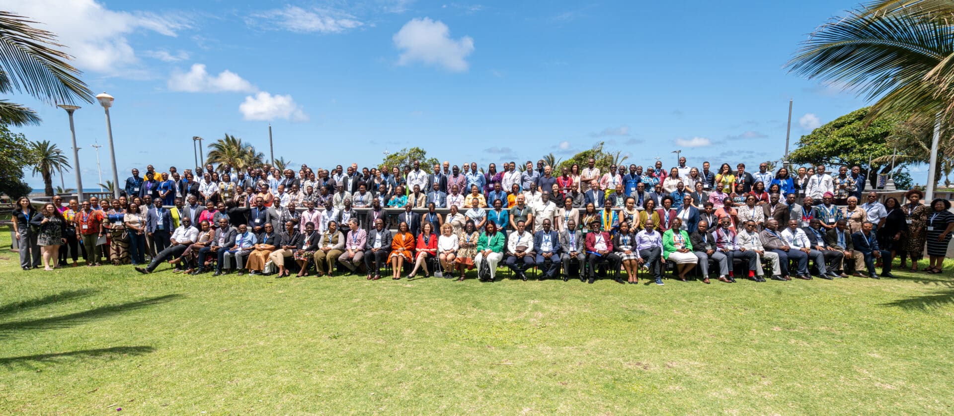 A large group of people pose for a photo outside on a sunny day, standing and sitting on chairs on a grassy lawn with palm trees and a blue sky in the background.