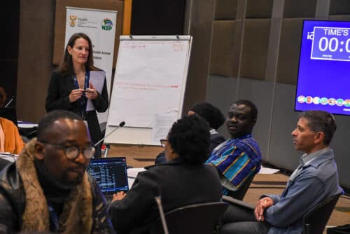 A woman stands and speaks near a flip chart in a conference room, while seated participants listen. A large digital timer is displayed on a screen in the background.