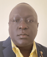 A person with short hair wearing a dark blazer over a light yellow collared shirt, facing the camera with a neutral expression against a plain light background.
