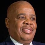 A man in a navy suit, light yellow striped tie, and white shirt is smiling. He has short hair and wears a red ribbon pin on his lapel, set against a dark background.