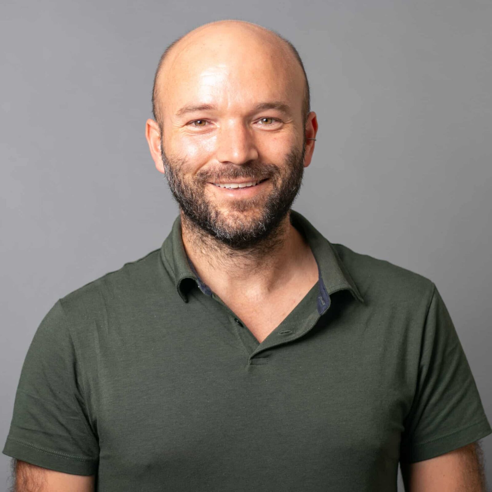 A smiling man with a beard and bald head wearing a short-sleeved, dark green polo shirt stands in front of a plain gray background.