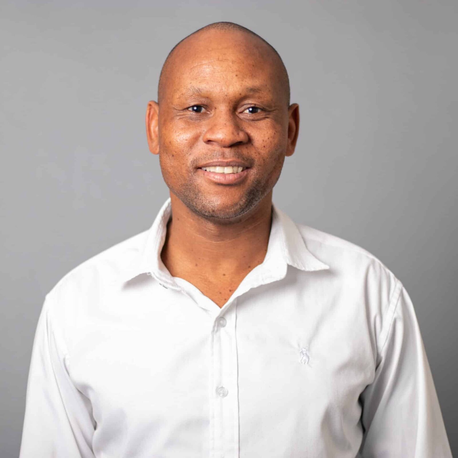 A man wearing a white collared shirt is smiling while standing against a plain gray background.