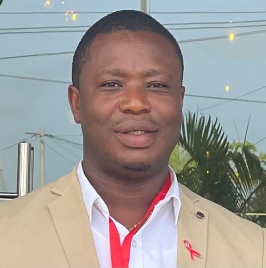 A man wearing a beige suit jacket, white shirt, and red lanyard stands outdoors, smiling. There is a red ribbon pinned to his jacket. Green plants and electrical wires are visible in the background.