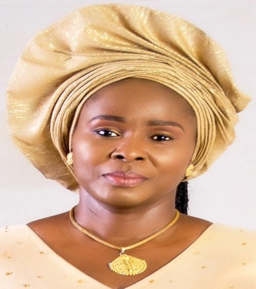 A woman wearing a gold headwrap and matching outfit, with gold earrings and a gold necklace, looking at the camera against a plain light background.