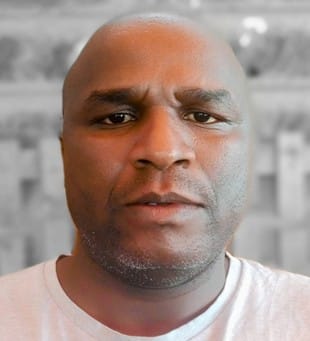 A man with a shaved head and slight stubble wears a light-colored shirt and looks directly at the camera, with a blurred, neutral background behind him.