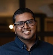 A man with short dark hair, glasses, and a beard is smiling. He is wearing a dark blue button-up shirt and standing indoors with softly lit, blurred background.