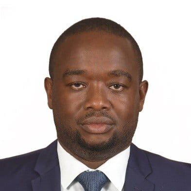 A man wearing a dark blue suit, white dress shirt, and blue tie is facing forward with a neutral expression against a plain white background.