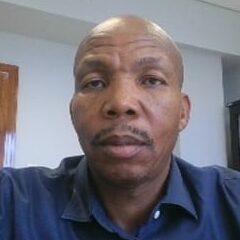 A man in a blue shirt sits at a desk in an office with shelves of binders and a wooden door in the background, looking directly at the camera.