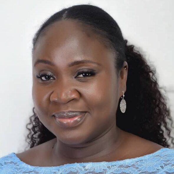 A woman with long, curly hair wears a light blue lace top and silver drop earrings, smiling gently against a plain white background.