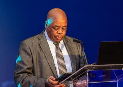 A man in a gray suit and polka dot tie stands at a clear podium, looking down at a tablet. There is a laptop on the podium and a microphone in front of him, with a blue background displaying partial text.