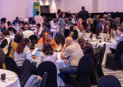 A large group of people sit in round tables engaged in discussions at a conference or workshop in a well-lit room. Some people use laptops, and a "YES You Can" sign is visible in the background.