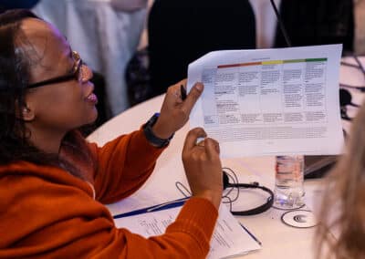 A woman in an orange sweater holds up a printed chart during a meeting, seated at a round table with documents, a water bottle, and headphones visible nearby.