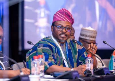 A man in traditional African attire and a pink cap speaks into a microphone at a conference table, surrounded by other participants and water bottles.