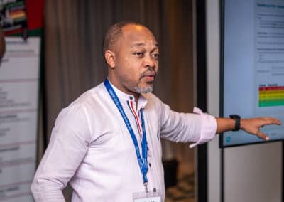 A man wearing a light pink shirt and blue lanyard gestures toward a large screen displaying a colorful dashboard, speaking to someone off camera in a conference setting.