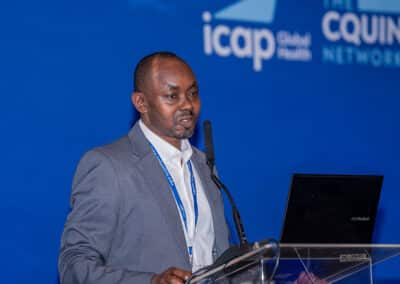 A man in a gray suit speaks at a podium with a microphone and a laptop, against a blue backdrop displaying the ICAP Global Health and CQUIN Network logos.