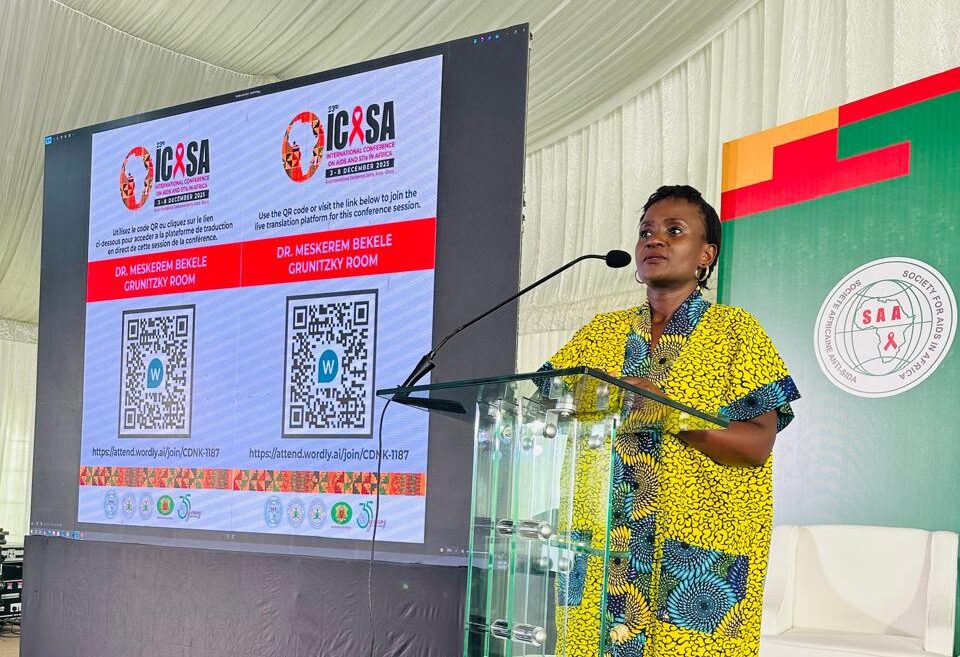 A woman in a yellow patterned dress stands at a clear podium, speaking into a microphone. Behind her is a large screen displaying conference information and QR codes, with a colorful event backdrop on the right.