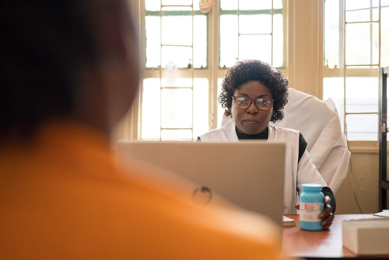 A healthcare professional wearing glasses sits at a desk with a laptop, talking to a patient. She holds a medication bottle, and sunlight streams through windows in the background.