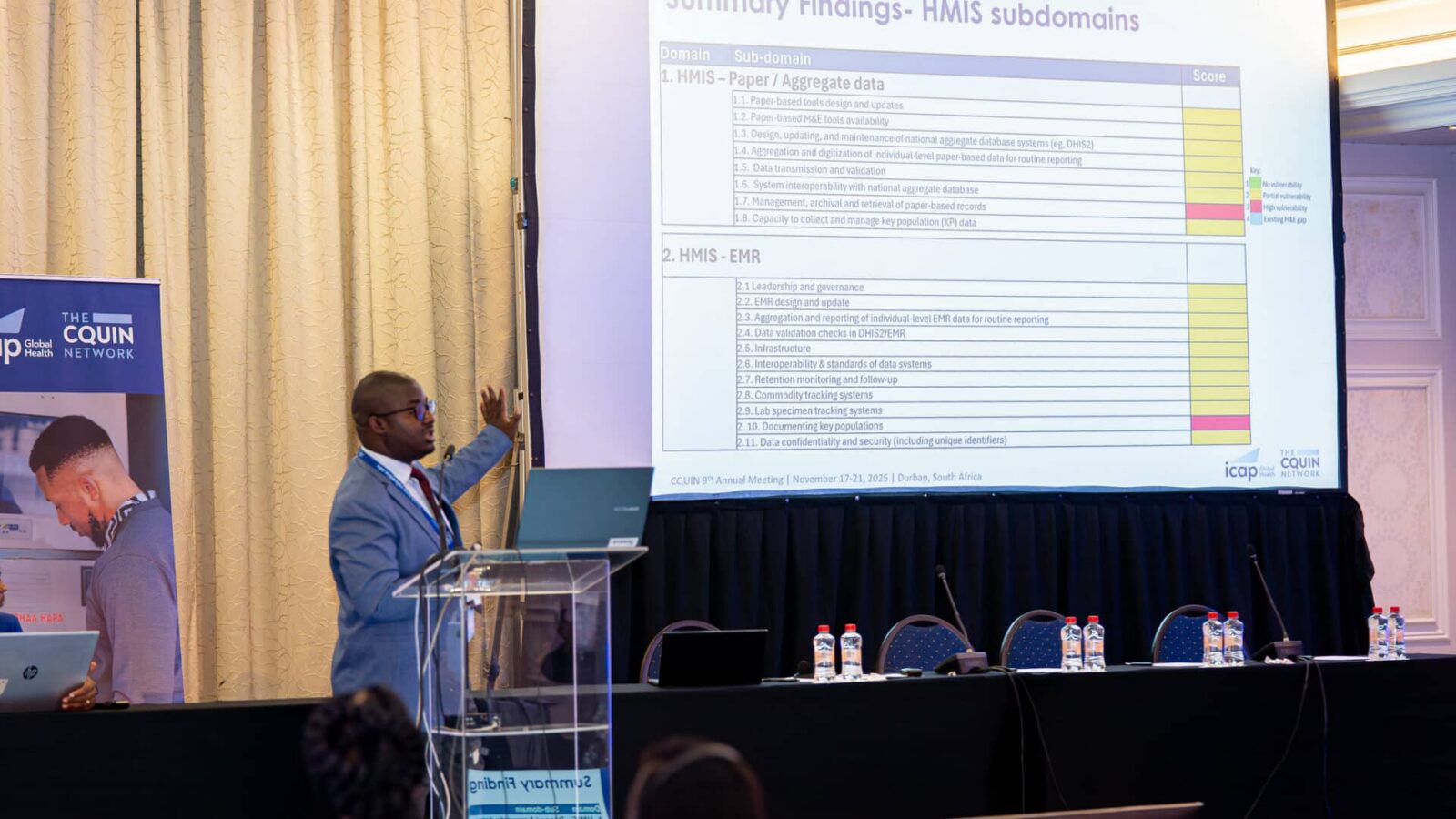 A man in a suit gives a presentation, pointing to a large projected slide with charts and text in a conference room. Several water bottles and microphones are on a table beside him.