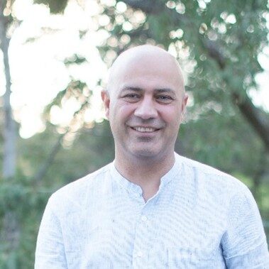 A smiling man with a shaved head and wearing a light-colored shirt stands outdoors, with greenery and trees softly blurred in the background.