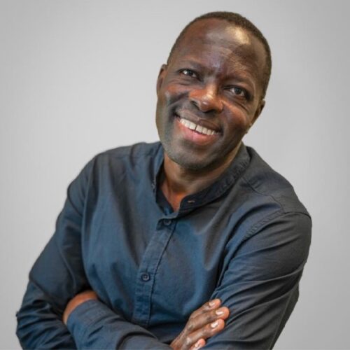 A man wearing a dark button-up shirt smiles warmly at the camera, with his arms folded across his chest, standing against a plain light gray background.