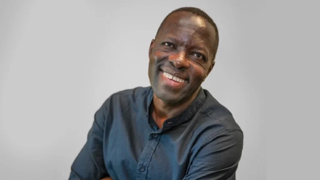 A man in a dark button-up shirt smiles warmly at the camera against a plain light grey background. He is standing with his arms crossed.