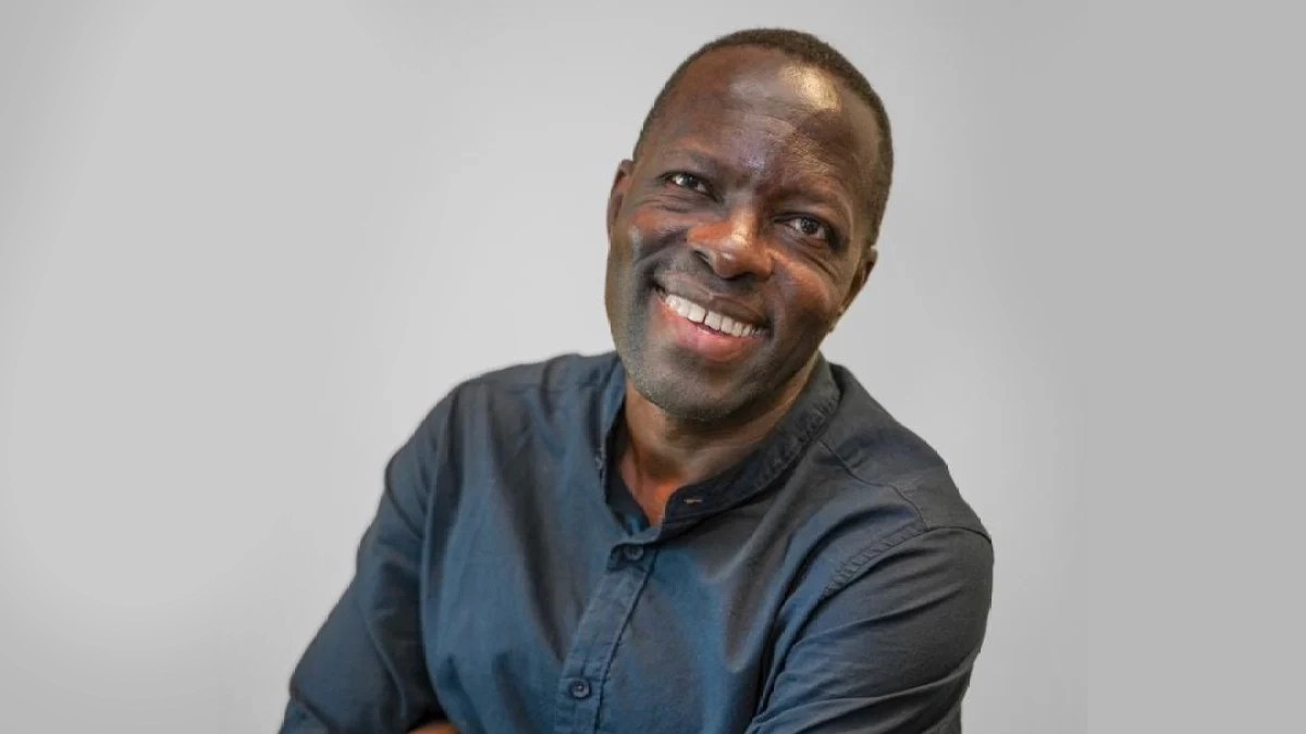 A man in a dark button-up shirt smiles warmly at the camera against a plain light grey background. He is standing with his arms crossed.