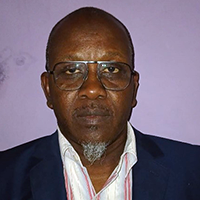 A man with glasses, a short grey goatee, and a serious expression is wearing a dark blazer over a collared shirt. He is standing in front of a light purple wall.