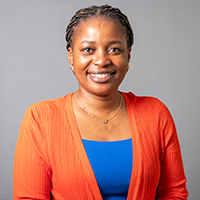A woman smiling at the camera, wearing a bright orange cardigan over a blue top, with a gray background. She has braided hair and is wearing a necklace and small earrings.