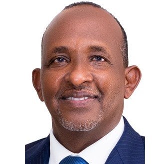 A middle-aged man with short hair and a trimmed beard, wearing a blue suit, white shirt, and blue tie, smiling at the camera against a plain white background.