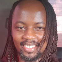 A smiling person with long, braided hair, wearing a dark shirt, is pictured indoors with a blurred background.