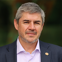 A man with short gray hair and a trimmed beard, wearing a dark blazer over a light purple shirt, stands outdoors with a blurred green background.