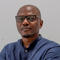 A man with dark skin and glasses, wearing a navy blue shirt, stands against a plain light gray background with his arms crossed, looking confidently at the camera.
