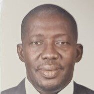 A man wearing a dark suit jacket, white shirt, and blue striped tie is facing the camera and smiling slightly against a plain light background.